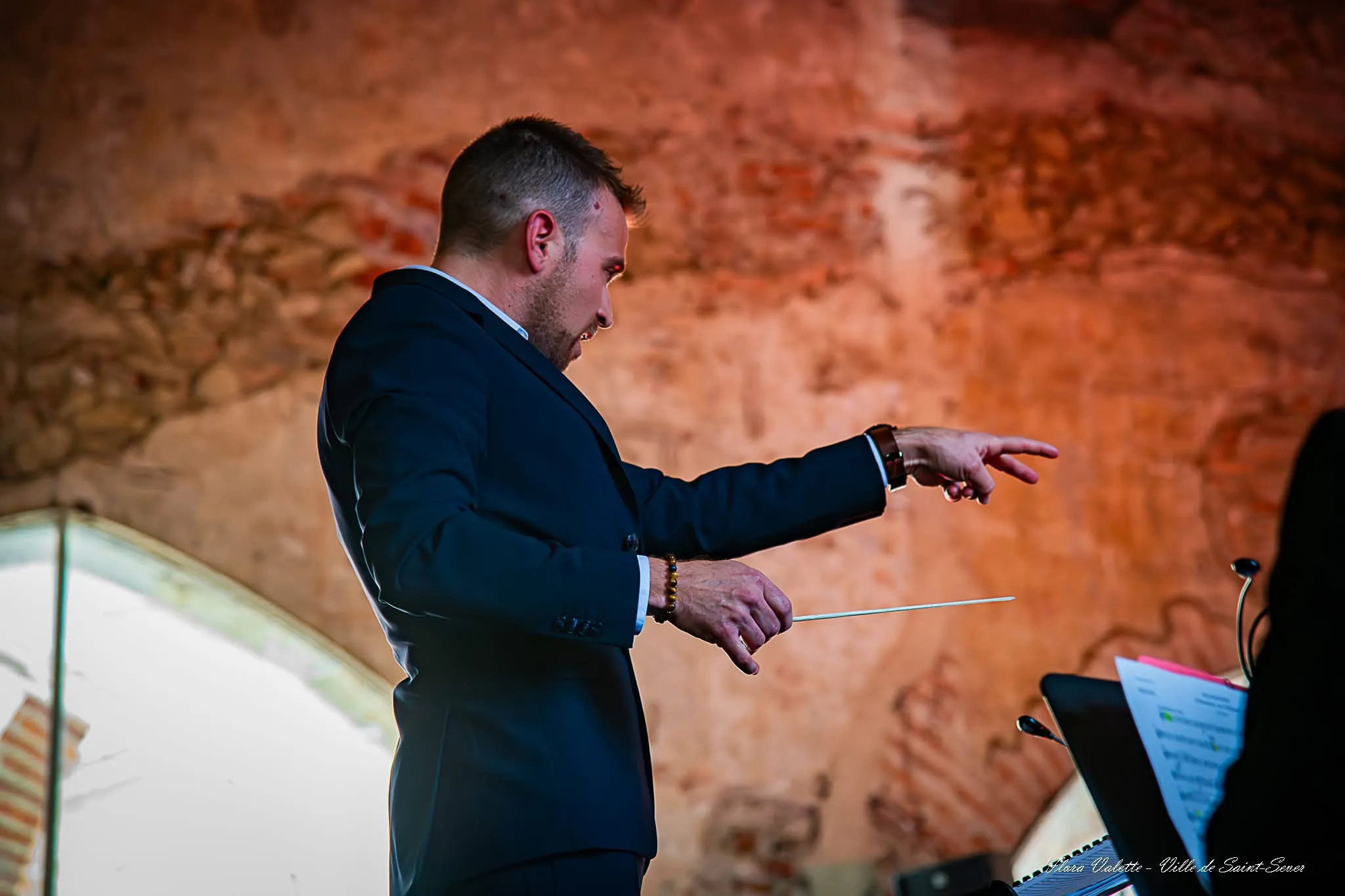 Julien Garcia dirigeant l'Orchestre d'Harmonie du Cap de Gascogne, baguette levée devant le mur de pierre du Cloître des Jacobins