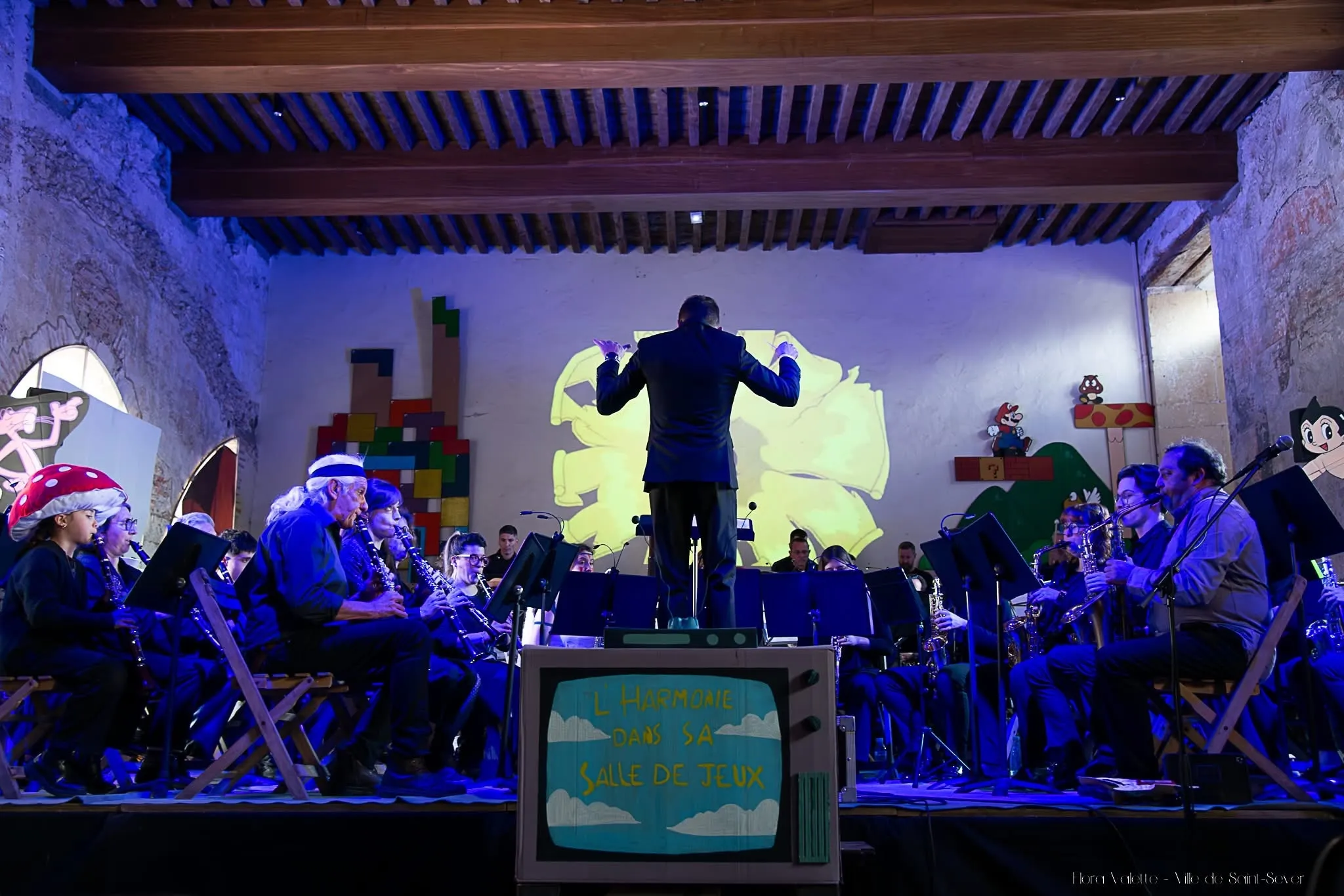 L'orchestre au complet sous une lumière bleue, entouré de décors Tetris, Mario et Astro Boy en carton, une télévision en carton « L'Harmonie dans sa salle de jeux » au premier plan
